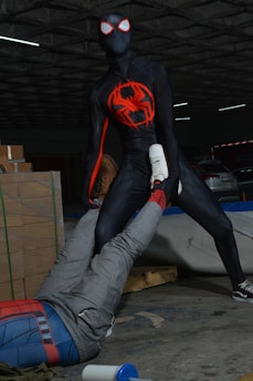 A person wearing a black spider-themed superhero costume with red accents stands in an assertive pose, holding the leg of another person dressed in a Spider-Man costume. The scene takes place in what appears to be a dimly lit warehouse with stacked cardboard boxes and a few visible parked cars.