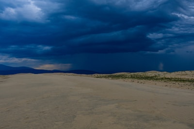 A vast desert scene with dramatic clouds casting shadows over rolling sand dunes.