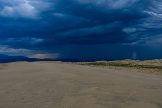 A vast desert scene with dramatic clouds casting shadows over rolling sand dunes.