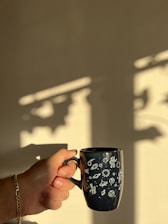 A hand is holding a dark mug decorated with white space-themed illustrations including planets, rockets, and astronauts. The background is a soft, blurred beige wall with dramatic shadows cast by a window frame, suggesting warm sunlight.