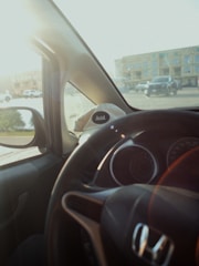 A view from inside a car showing an outside parking lot with several vehicles and a building in the background. The car's steering wheel is visible, and there is a hat with the word 'Avid.' resting on the dashboard.