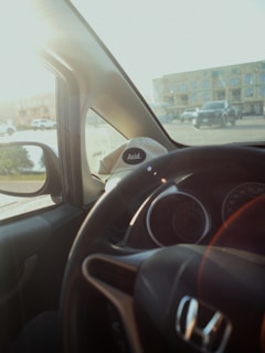 A view from inside a car showing an outside parking lot with several vehicles and a building in the background. The car's steering wheel is visible, and there is a hat with the word 'Avid.' resting on the dashboard.
