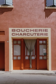 A storefront with a rustic red and beige color scheme, featuring a closed door with a tied curtain. The sign above the entrance reads 'Boucherie Charcuterie' and 'Traiteur' is written vertically on the sides. The building has a traditional appearance with small shutters at the top.