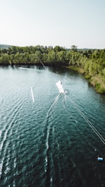 Aerial view of a person wakeboarding on a lake surrounded by lush greenery. The water is a deep blue-green, and the wake from the board creates patterns on the surface. A dense line of trees borders the far side of the lake, giving a sense of seclusion.