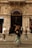 An ornate, historic building entrance with the sign 'Grand Pera' above the archway is bustling with pedestrian activity. The building facade features detailed architecture, including columns and decorative elements. People are walking across the street in front of the entrance, some blurred due to movement, while shops line the sides of the entrance.