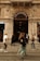 An ornate, historic building entrance with the sign 'Grand Pera' above the archway is bustling with pedestrian activity. The building facade features detailed architecture, including columns and decorative elements. People are walking across the street in front of the entrance, some blurred due to movement, while shops line the sides of the entrance.