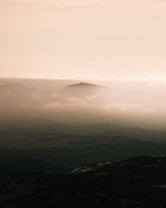 A tranquil landscape with layers of mountains shrouded in mist, creating a serene and ethereal atmosphere. The soft beige tones dominate the sky, while darker shades define the distant hills.