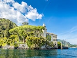 A picturesque villa stands on a lush, green hillside by the water. The building is surrounded by tall, manicured trees and vibrant greenery. The sky above is bright blue with a few scattered clouds, and the water reflects the serene and clear atmosphere. The scene is tranquil and beautifully maintained, suggesting a peaceful, natural environment.