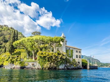 A picturesque villa stands on a lush, green hillside by the water. The building is surrounded by tall, manicured trees and vibrant greenery. The sky above is bright blue with a few scattered clouds, and the water reflects the serene and clear atmosphere. The scene is tranquil and beautifully maintained, suggesting a peaceful, natural environment.