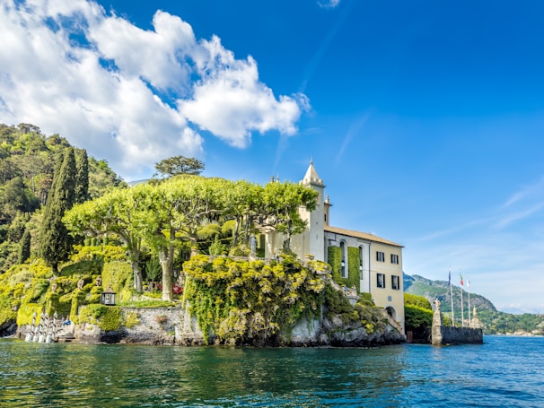 A picturesque villa stands on a lush, green hillside by the water. The building is surrounded by tall, manicured trees and vibrant greenery. The sky above is bright blue with a few scattered clouds, and the water reflects the serene and clear atmosphere. The scene is tranquil and beautifully maintained, suggesting a peaceful, natural environment.