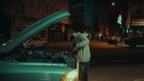 A person is working under the open hood of a car on a dimly lit street at night. Buildings and streetlights in the background create an urban setting, with a few other parked cars visible along the road. The person is wearing a warm hat and jacket, suggesting cool weather.