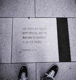 An engraved stone tile on the ground displays a quote from Proverbs 31:26, surrounded by textured concrete tiles. A pair of black sneakers with white laces and soles is visible at the bottom of the image, indicating that the photo was taken from a first-person perspective.