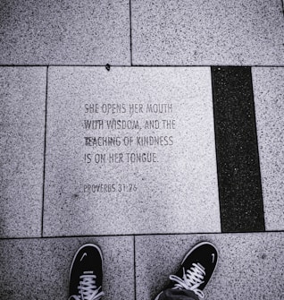 An engraved stone tile on the ground displays a quote from Proverbs 31:26, surrounded by textured concrete tiles. A pair of black sneakers with white laces and soles is visible at the bottom of the image, indicating that the photo was taken from a first-person perspective.