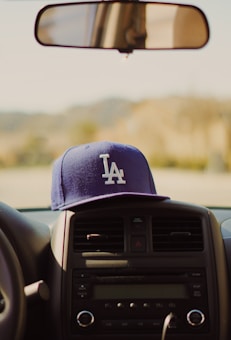 A blue baseball cap with the letters 'LA' on the front is placed on the dashboard of a car, directly beneath the rearview mirror. The interior of the car is partially visible, focusing on the radio and air vents.