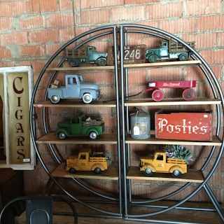 A shelf against a brick wall displays vintage toy trucks in various colors, a red toy wagon, and a tin container. Signs reading 'Cigars' and 'Posties' add to the nostalgic feel.
