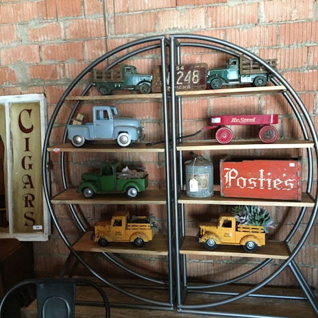 A shelf against a brick wall displays vintage toy trucks in various colors, a red toy wagon, and a tin container. Signs reading 'Cigars' and 'Posties' add to the nostalgic feel.