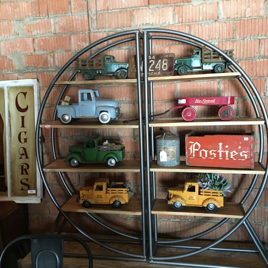 A shelf against a brick wall displays vintage toy trucks in various colors, a red toy wagon, and a tin container. Signs reading 'Cigars' and 'Posties' add to the nostalgic feel.