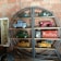 A shelf against a brick wall displays vintage toy trucks in various colors, a red toy wagon, and a tin container. Signs reading 'Cigars' and 'Posties' add to the nostalgic feel.