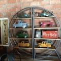 A shelf against a brick wall displays vintage toy trucks in various colors, a red toy wagon, and a tin container. Signs reading 'Cigars' and 'Posties' add to the nostalgic feel.