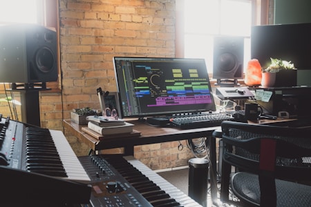 A music production studio featuring a large monitor displaying audio editing software surrounded by various equipment such as a keyboard, speakers, and controllers. Natural light enters through the window, illuminating the brick wall and a desk cluttered with books, headphones, and small plants, including a glowing salt lamp.