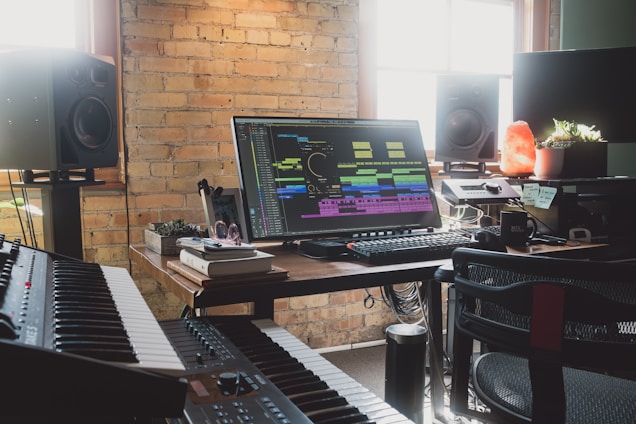 A music production studio featuring a large monitor displaying audio editing software surrounded by various equipment such as a keyboard, speakers, and controllers. Natural light enters through the window, illuminating the brick wall and a desk cluttered with books, headphones, and small plants, including a glowing salt lamp.