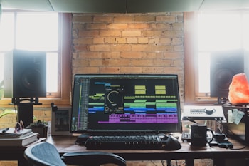 A music production workspace featuring a large monitor displaying audio editing software. The desk is surrounded by two large studio speakers. Nearby, there is a keyboard, a mouse, and some personal items like a mug and a Himalayan salt lamp. The background is a brick wall with windows on either side, allowing natural light to fill the room.