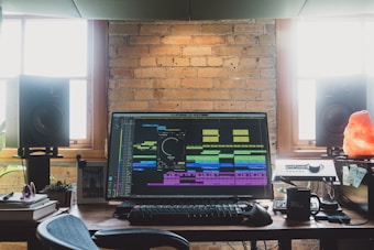 A music production workspace featuring a large monitor displaying audio editing software. The desk is surrounded by two large studio speakers. Nearby, there is a keyboard, a mouse, and some personal items like a mug and a Himalayan salt lamp. The background is a brick wall with windows on either side, allowing natural light to fill the room.