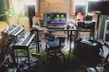 A home music studio setup with digital audio workstation software displayed on a computer monitor. The room contains synthesizers, a keyboard, guitars, studio monitor speakers, effect pedals, and an amplifier. Natural light comes through the window, and there are various decorative elements like a potted plant and a Himalayan salt lamp.