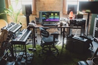 A home music studio setup with digital audio workstation software displayed on a computer monitor. The room contains synthesizers, a keyboard, guitars, studio monitor speakers, effect pedals, and an amplifier. Natural light comes through the window, and there are various decorative elements like a potted plant and a Himalayan salt lamp.