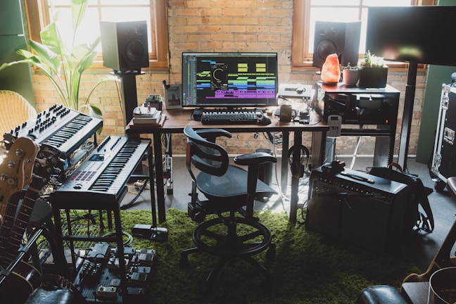 A home music studio setup with digital audio workstation software displayed on a computer monitor. The room contains synthesizers, a keyboard, guitars, studio monitor speakers, effect pedals, and an amplifier. Natural light comes through the window, and there are various decorative elements like a potted plant and a Himalayan salt lamp.