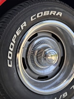 Close-up of a car tire with the words 'COOPER COBRA' prominently displayed in white letters on the black rubber surface. The tire is mounted on a metal rim with intricate, radial design and a shiny, reflective central hubcap.