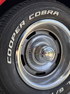 Close-up of a car tire with the words 'COOPER COBRA' prominently displayed in white letters on the black rubber surface. The tire is mounted on a metal rim with intricate, radial design and a shiny, reflective central hubcap.