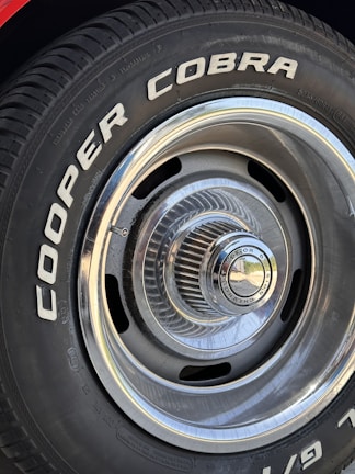 Close-up of a car tire with the words 'COOPER COBRA' prominently displayed in white letters on the black rubber surface. The tire is mounted on a metal rim with intricate, radial design and a shiny, reflective central hubcap.
