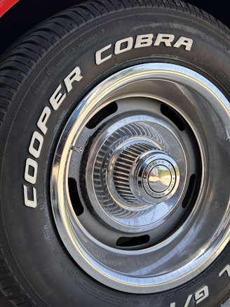 Close-up of a car tire with the words 'COOPER COBRA' prominently displayed in white letters on the black rubber surface. The tire is mounted on a metal rim with intricate, radial design and a shiny, reflective central hubcap.