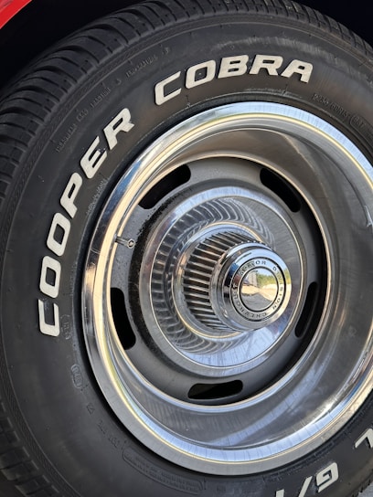 Close-up of a car tire with the words 'COOPER COBRA' prominently displayed in white letters on the black rubber surface. The tire is mounted on a metal rim with intricate, radial design and a shiny, reflective central hubcap.