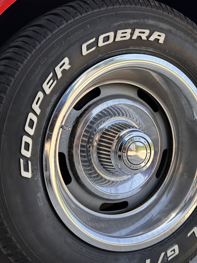 Close-up of a car tire with the words 'COOPER COBRA' prominently displayed in white letters on the black rubber surface. The tire is mounted on a metal rim with intricate, radial design and a shiny, reflective central hubcap.