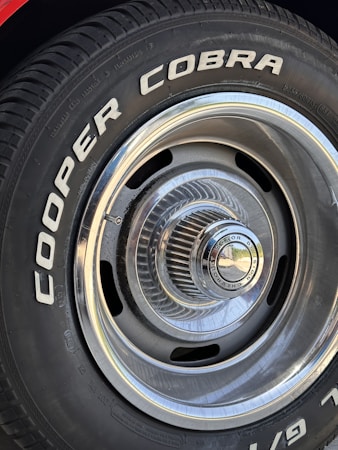 Close-up of a car tire with the words 'COOPER COBRA' prominently displayed in white letters on the black rubber surface. The tire is mounted on a metal rim with intricate, radial design and a shiny, reflective central hubcap.