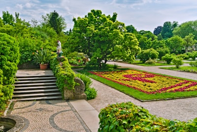 A well-maintained garden landscape featuring neatly arranged flower beds with geometric patterns, surrounded by lush green trees and shrubs. A patterned stone walkway leads up stone steps, bordered by vibrant green ivy. A decorative statue or sculpture is placed on a pedestal next to a potted plant. The background includes various tall trees under a partly cloudy sky.