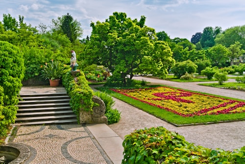 A well-maintained garden landscape featuring neatly arranged flower beds with geometric patterns, surrounded by lush green trees and shrubs. A patterned stone walkway leads up stone steps, bordered by vibrant green ivy. A decorative statue or sculpture is placed on a pedestal next to a potted plant. The background includes various tall trees under a partly cloudy sky.