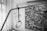 A hospital lifting device is positioned next to a bed, with its handle hanging in the foreground. In the background, a monochrome painting features abstract figures holding an object, surrounded by flowing shapes and motifs reminiscent of fish.