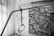 A hospital lifting device is positioned next to a bed, with its handle hanging in the foreground. In the background, a monochrome painting features abstract figures holding an object, surrounded by flowing shapes and motifs reminiscent of fish.