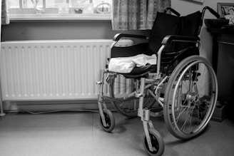 A black and white image of a wheelchair positioned next to a radiator and underneath a window with curtains. The wheelchair is empty, and a towel or cloth is draped over the seat. The setting appears to be a room with a wooden piece of furniture on the right.