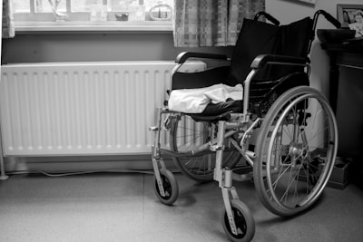 A black and white image of a wheelchair positioned next to a radiator and underneath a window with curtains. The wheelchair is empty, and a towel or cloth is draped over the seat. The setting appears to be a room with a wooden piece of furniture on the right.