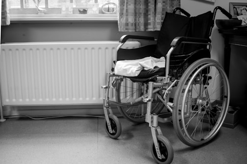 A black and white image of a wheelchair positioned next to a radiator and underneath a window with curtains. The wheelchair is empty, and a towel or cloth is draped over the seat. The setting appears to be a room with a wooden piece of furniture on the right.