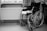 A black and white image of a wheelchair positioned next to a radiator and underneath a window with curtains. The wheelchair is empty, and a towel or cloth is draped over the seat. The setting appears to be a room with a wooden piece of furniture on the right.