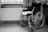 A black and white image of a wheelchair positioned next to a radiator and underneath a window with curtains. The wheelchair is empty, and a towel or cloth is draped over the seat. The setting appears to be a room with a wooden piece of furniture on the right.