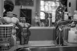 A bathroom countertop with a metal faucet, two uniquely shaped perfume bottles resembling human torsos, a paper cup with a checkered pattern, and soap dispensers in the background. The scene is monochromatic, focusing on reflections and textures.