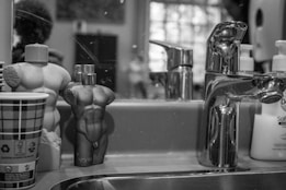 A bathroom countertop with a metal faucet, two uniquely shaped perfume bottles resembling human torsos, a paper cup with a checkered pattern, and soap dispensers in the background. The scene is monochromatic, focusing on reflections and textures.