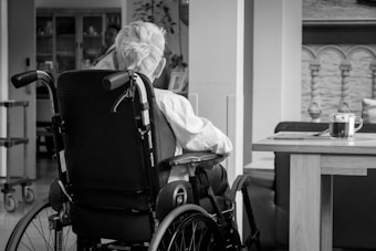 An older person with white hair is sitting in a wheelchair facing away in a cozy indoor setting. A small wooden table nearby holds a glass mug of tea placed on a coaster. The background features some home furnishings and decorative elements.