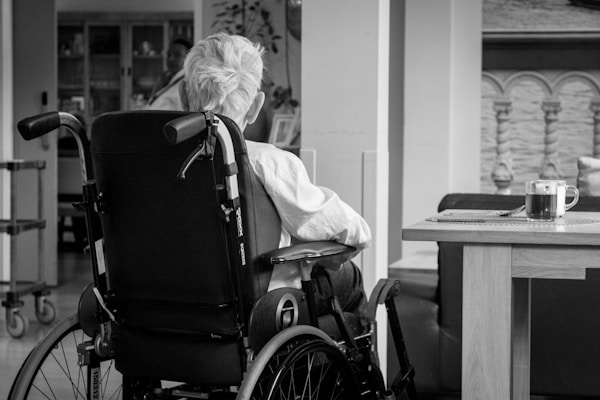 An older person with white hair is sitting in a wheelchair facing away in a cozy indoor setting. A small wooden table nearby holds a glass mug of tea placed on a coaster. The background features some home furnishings and decorative elements.